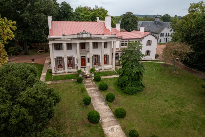 Aerial view of a historic mansion with red roof and columns surrounded by trees and garden pathways.