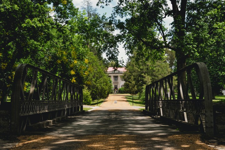 View of a metal bridge leading to a white house surrounded by trees.