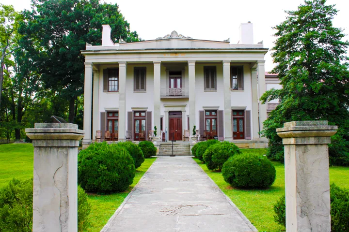 Large, historic white mansion with columns, surrounded by trees and bushes.