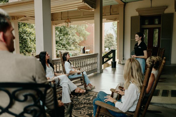 People sitting on a porch listening to someone talk.