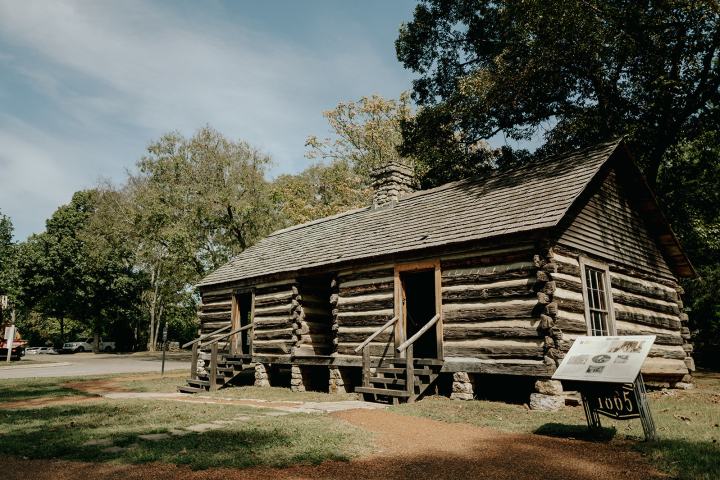 Rustic log cabin with a gabled roof and two porches in a wooded area.