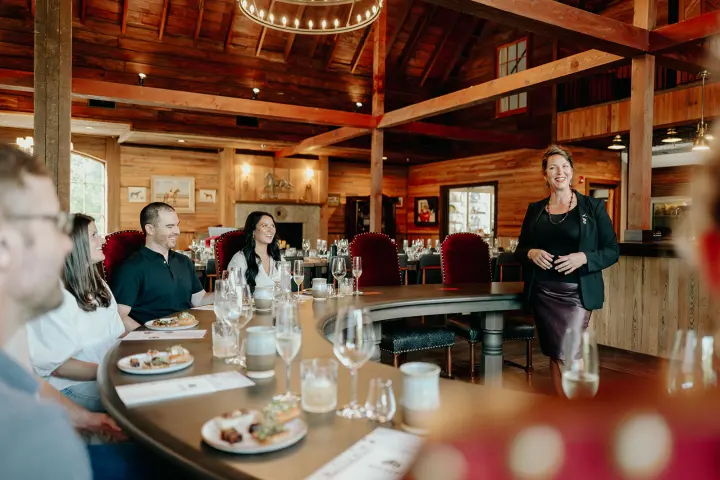 Woman speaking to seated people in a rustic dining setting.
