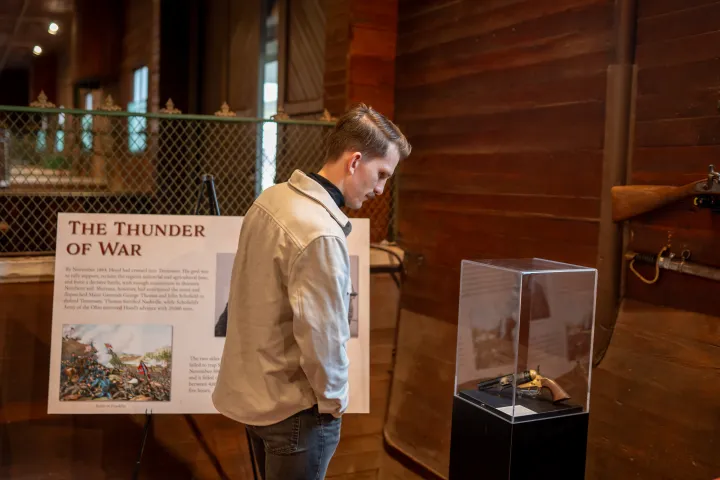 Man observing war exhibit, titled 'The Thunder of War', in museum with a rifle on the wall.