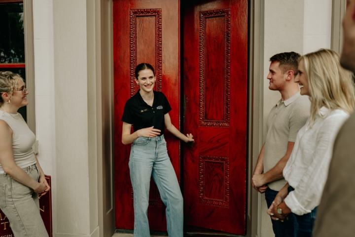 A group of people stand outside a building with red doors, smiling and chatting.