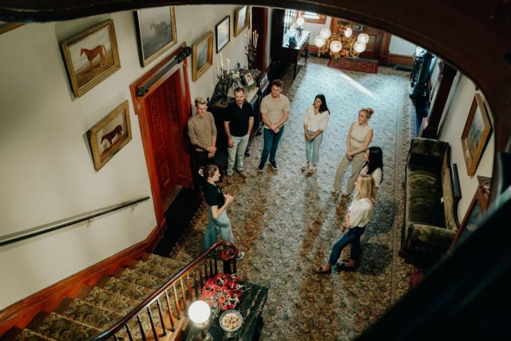 Group of people standing in a large, ornate hallway with paintings on the walls.