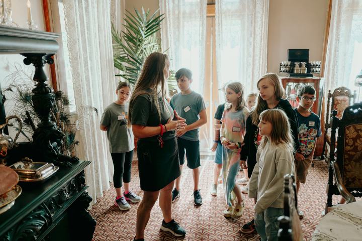 Woman giving a tour to a group of children in a room filled with antique furniture.