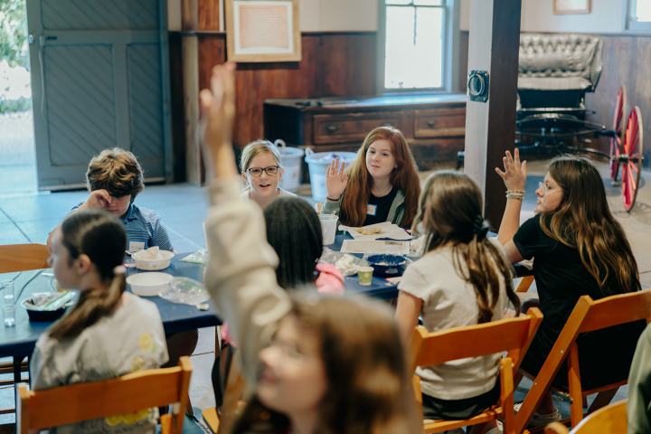 Children sitting around a table, some raising hands, in a rustic room with a barn-like interior.
