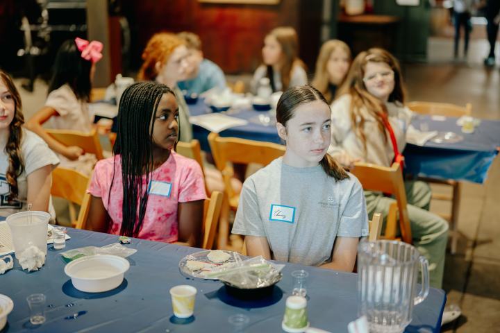 Group of children sitting at a table with art supplies, wearing name tags, in a classroom setting.