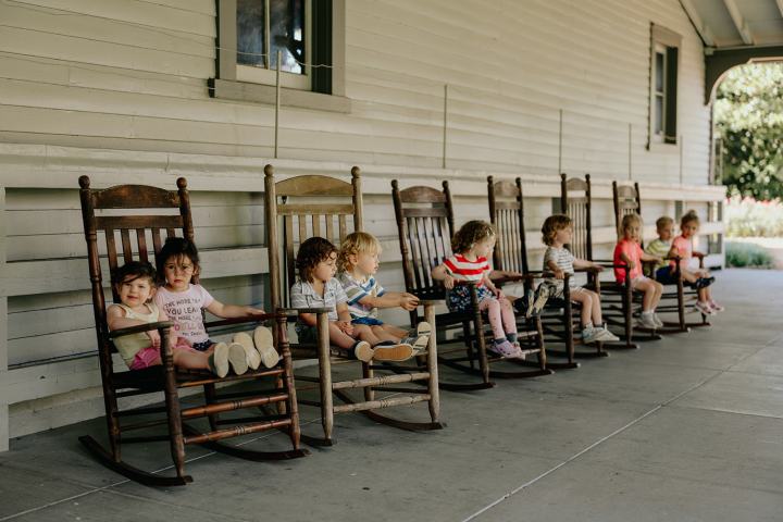 Eight children sitting in a row of wooden rocking chairs on a porch.