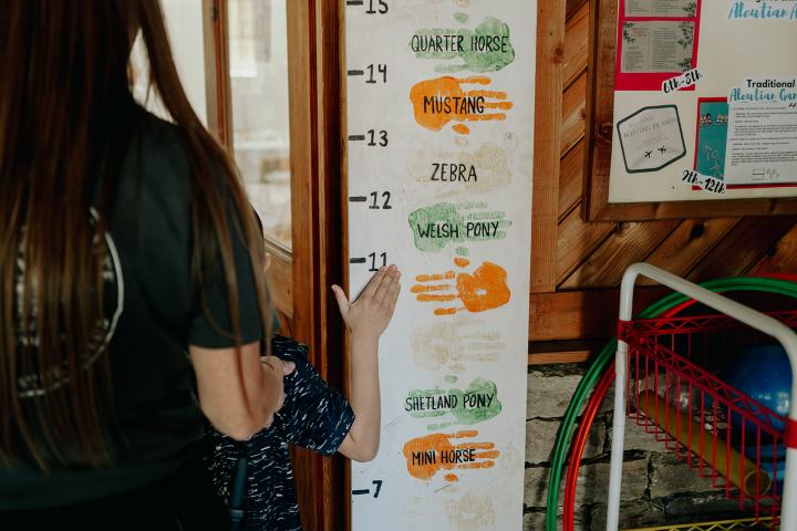 Child measuring height against a chart with handprints and equine names, supervised by an adult.