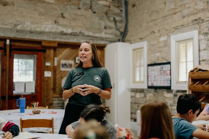 Woman speaking to children in a rustic classroom setting.