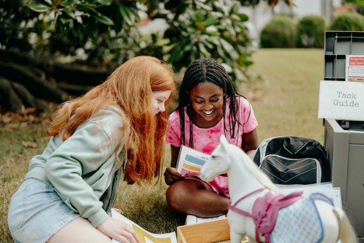 Two girls sitting on grass looking at papers near a toy horse and a tack guide.
