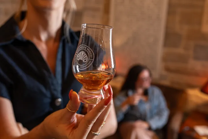 Person holding a glass with amber liquid, blurred person in background.