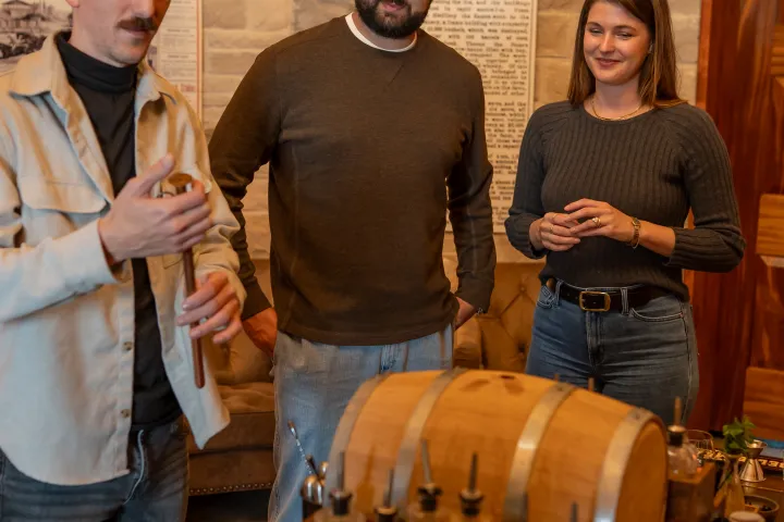 Three people standing around a bar setup with bottles and small barrels.