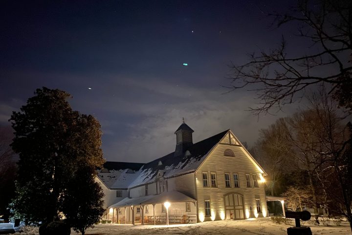 A lit barn in a snowy landscape under a cloudy night sky with trees.