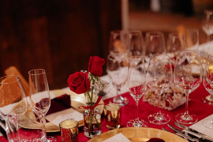 Elegant dining table with red roses, gold plates, and crystal glasses set on a red table runner.