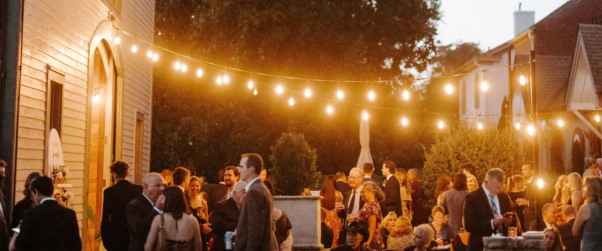 Outdoor evening gathering with string lights and a crowd in formal attire at a brick-building venue.
