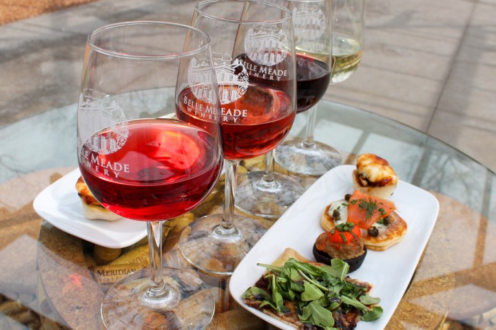 Four wine glasses and assorted appetizers on a glass table outdoors.