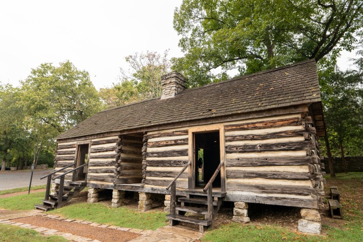 Old log cabin with stone foundation and two entrances, surrounded by trees.