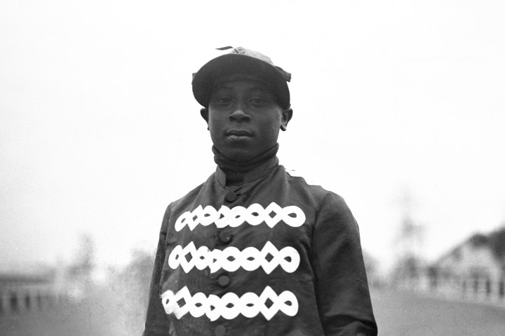 Black and white photo of a person in a jockey uniform with ornate buttons, standing outdoors.