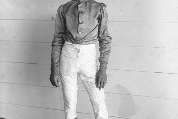 Black and white photo of a jockey in uniform standing against a wooden wall.