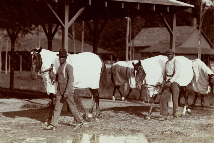 Two men with horses wearing blankets near a wooden structure.