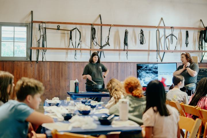People in a classroom with tools on wall, presenter talks, students sit at tables.