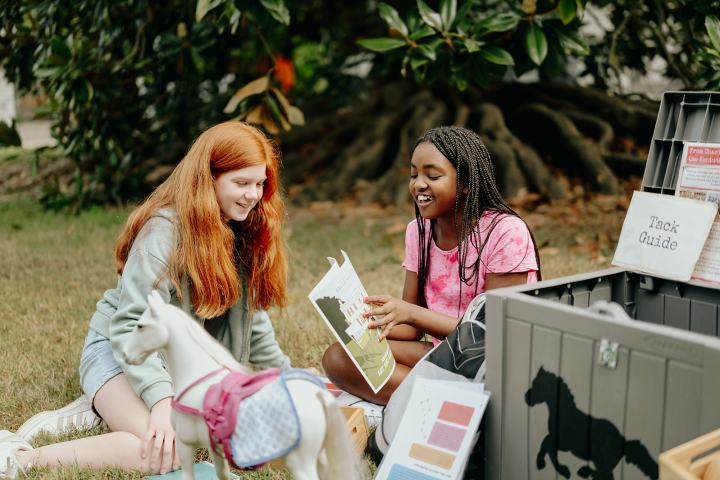 Two girls outdoors reading a book next to a toy horse.