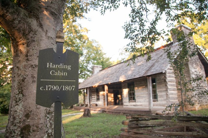 Historic log cabin with 'Harding Cabin c.1790/1807' sign, surrounded by trees and wooden fence.