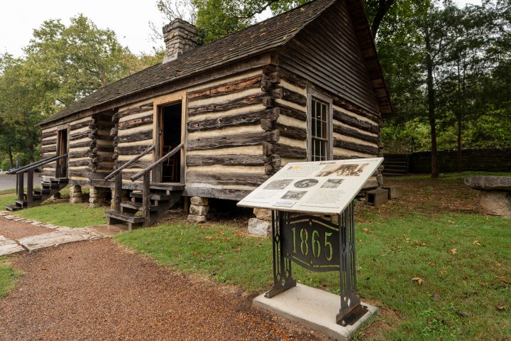 Historic log cabin with informational sign and trees in background.