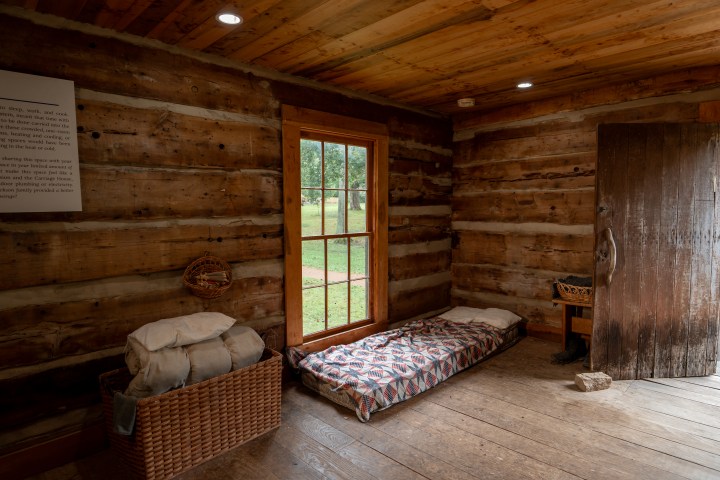Rustic log cabin interior with wooden floors, a simple bed, window, and a basket with blankets.