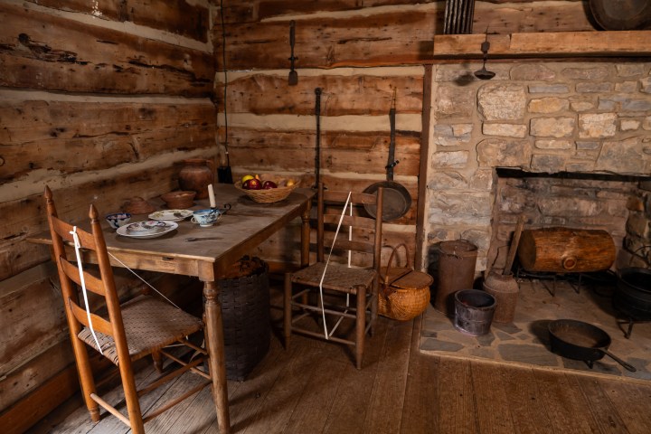 Rustic cabin interior with wooden table, chairs, and stone fireplace.