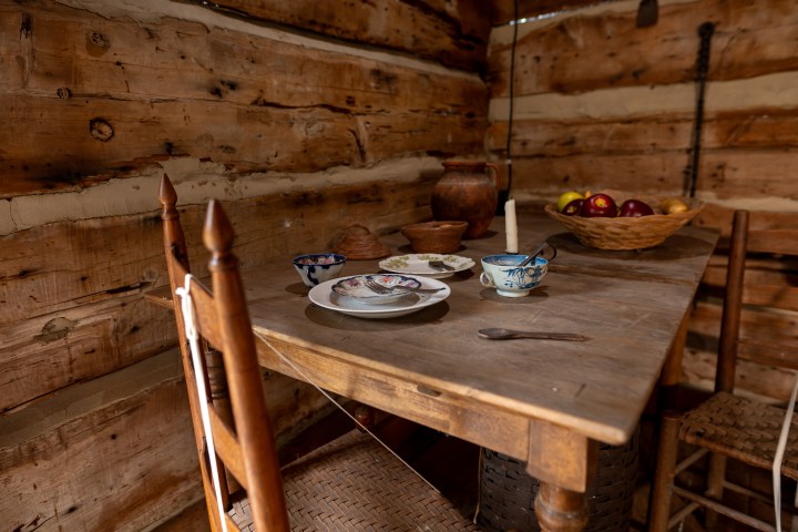 Rustic wooden table with dishes, cups, and fruit bowl in a log cabin.