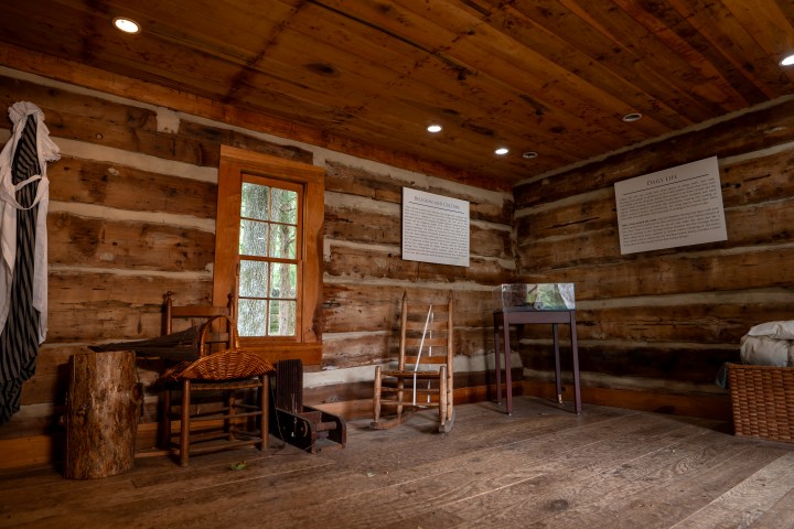 Rustic log cabin interior with chairs, a spinning wheel, and natural wood walls.