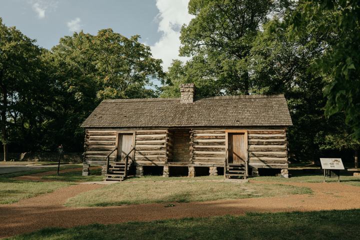 Historic log cabin with two doors, surrounded by trees.