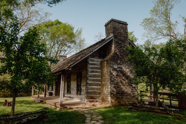 Rustic log cabin with stone chimney, surrounded by trees and grass.