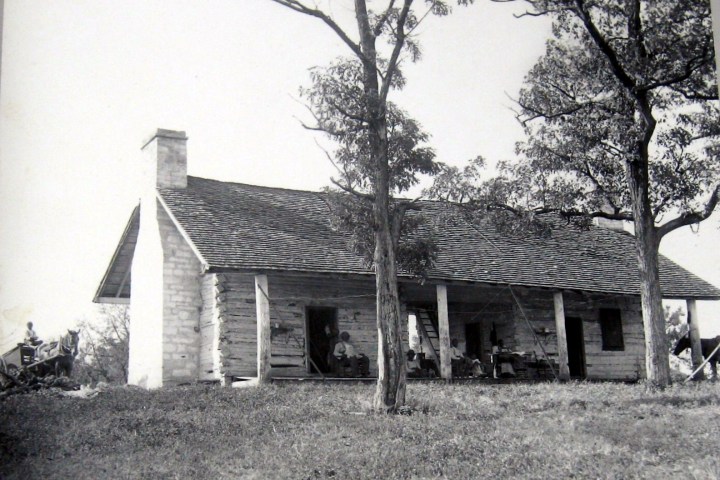 Old log cabin with a porch, surrounded by trees.