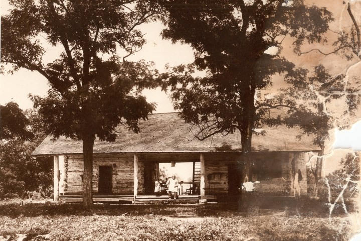 Sepia photo of an old log cabin with a porch, two large trees, and two people standing in front.