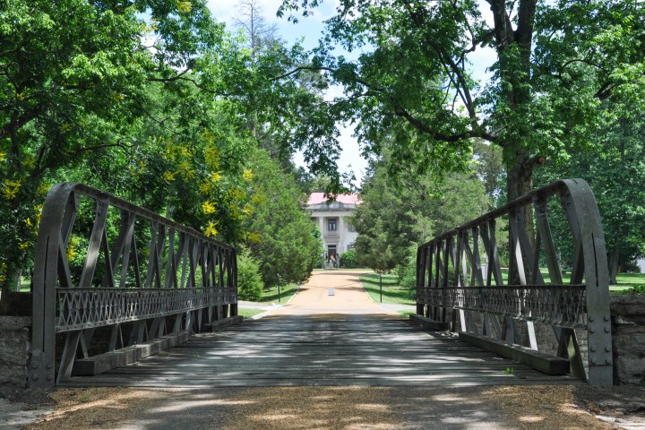 Metal bridge leading to a historic house surrounded by trees.