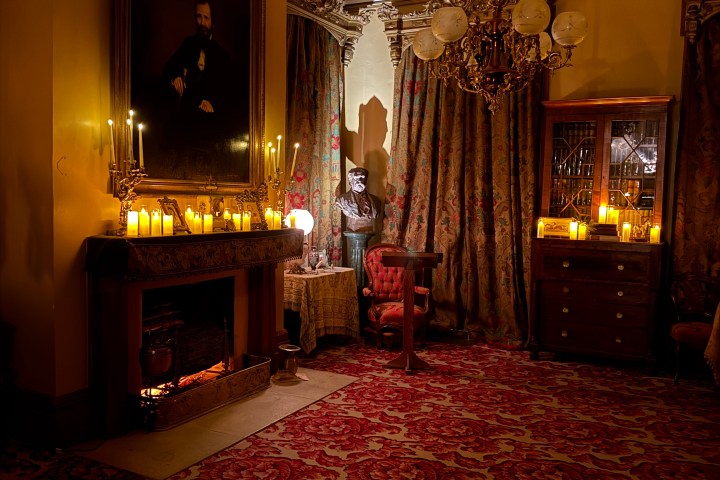 Dimly lit vintage room with portrait, fireplace, candles, and ornate red carpet.